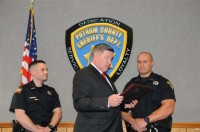 Sheriff Smith reading the award citations aloud as he is flanked by Deputy Blessing on the viewer’s left and Deputy Hunsberger on the right.  